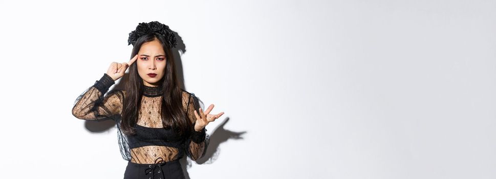 Frustrated asian woman in halloween costume arguing and complaining, scolding for something stupid, pointing finger at head and grimacing, standing over white background.