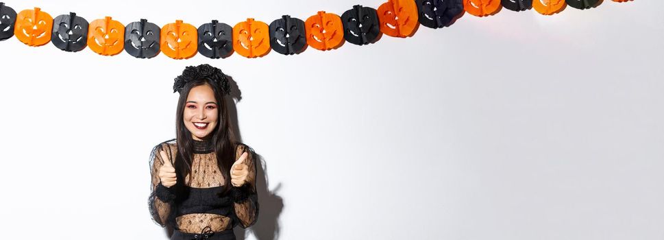 Image of happy and satisfied asian female in witch costume showing super, thumbs-up gesture and smiling pleased, standing against pumpkins banner.