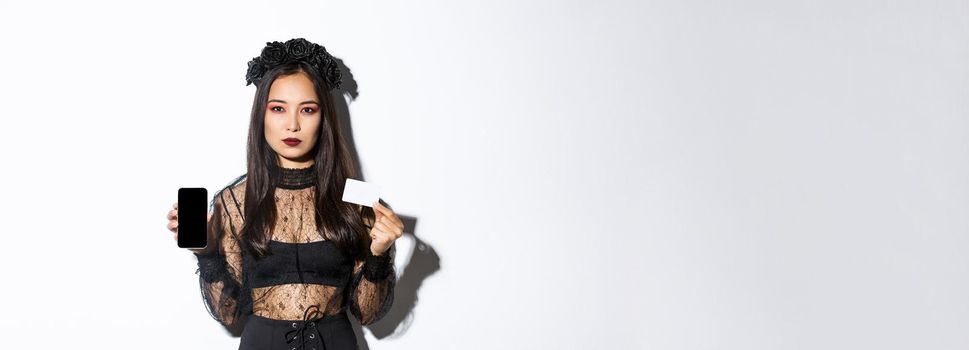 Serious-looking elegant young asian woman in gothic dress and black wreath showing credit card and mobile phone screen, celebrating halloween.