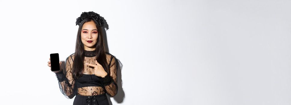 Young elegant woman in gothic dress and black wreath pointng finger at smartphone screen with pleased smile on her face, standing over white background.