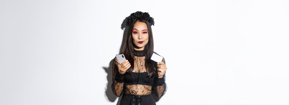 Sassy young woman looking thoughtful, holding credit card and mobile phone, shopping in internet, standing over white background.