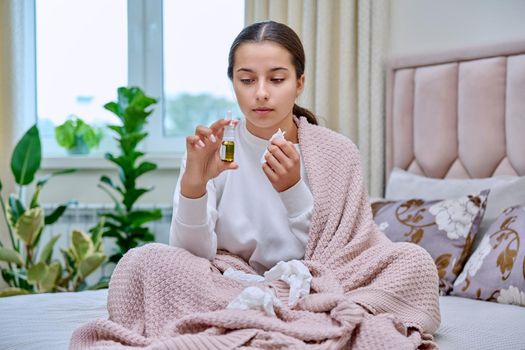 Young teenage female sitting in bed with runny nose spray drops, sneezing into tissues, burying her nose. Autumn winter, rhinitis runny nose stuffy nose, treatment, flu season