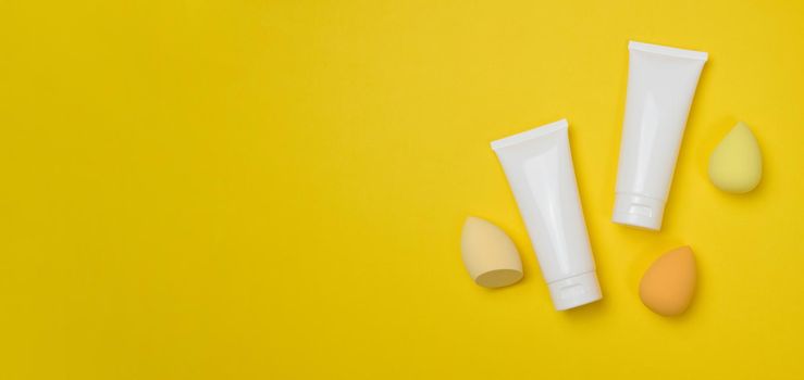 White plastic tubes for cream, gel and other cosmetics and sponges on a yellow background, top view