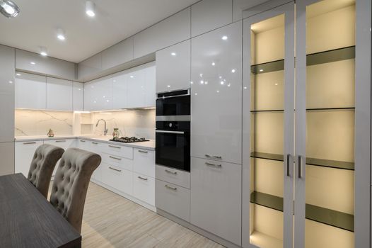 Dining room table and chairs, white spacious furnished kitchen interior with oven and large LED enlightened sideboard at right side