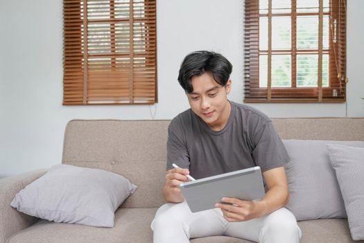 Handsome Asian man using digital tablet while headphones sitting on sofa at home.