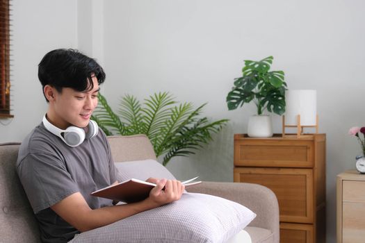 Handsome Asian man relaxing on the sofa at home with a book and headphones.
