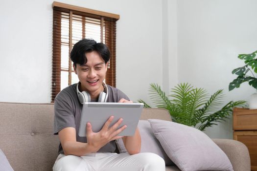Handsome Asian man using digital tablet while headphones sitting on sofa at home.