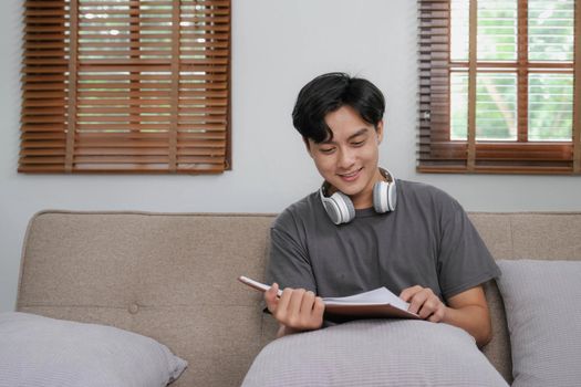 Handsome Asian man relaxing on the sofa at home with a book and headphones.