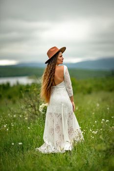 A middle-aged woman in a white dress and brown hat stands on a green field and holds a basket in her hands with a large bouquet of daisies. In the background there are mountains and a lake