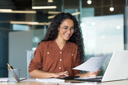 Young successful businesswoman behind paperwork, Hispanic woman working with documents and contracts inside modern office building, female worker using laptop, wearing glasses and curly hair smiling.
