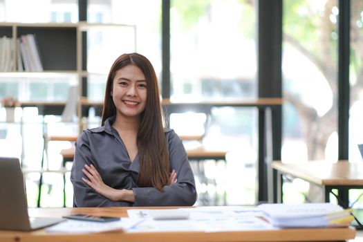 Confident young Asian business woman sitting with arms crossed smiling looking at camera in the office..