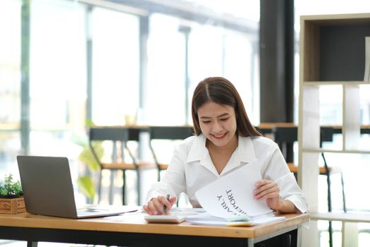 Smiling beautiful Asian businesswoman analyzing chart and graph showing changes on the market and use calculator at office..