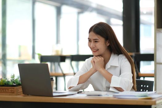 Beautiful young Asian businesswoman is smiling at her desk and taking notes with computer laptop on her desk, enjoying work..