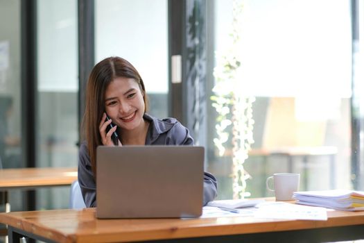 Young Asian businesswoman sit with her laptop computer happily talking on the phone with her customer explaining the detail in the office..