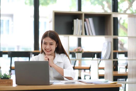 Beautiful young Asian businesswoman is smiling at her desk and taking notes with computer laptop on her desk, enjoying work..