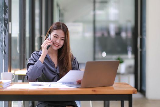 Young Asian businesswoman sit with her laptop computer happily talking on the phone with her customer explaining the detail in the office..