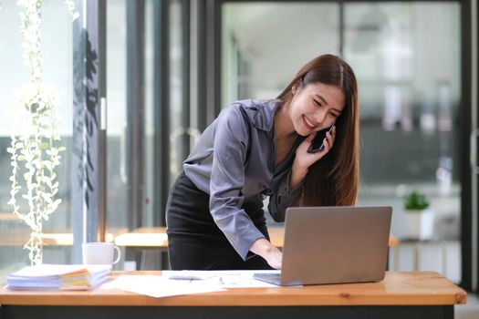 Happy young Asian businesswoman standing using smartphone and laptop computer at office..