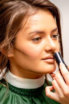 Beautiful young brunette woman receiving makeup with stick concealer on her face in a beauty salon