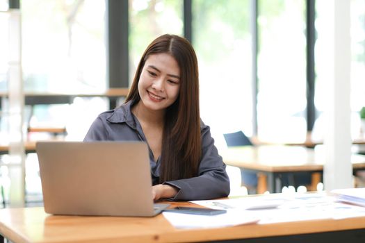 Beautiful Asian business woman using her laptop to work and enjoy working, taking note, reviewing assignment and smiling in the office..