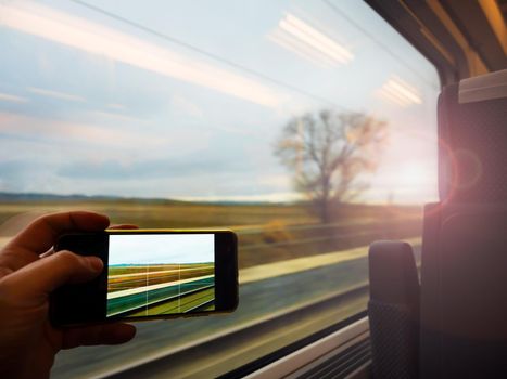 Subjective shoot of one hand taking a picture of the landscape through the window of the moving train with a smart phone. Dynamic image due to motion blur. Sunlight creates soft and romantic effect