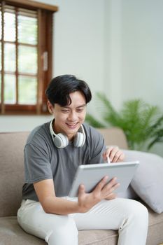 A portrait of a young man showing a smiling face while relaxing, using a tablet computer and a pair of headphones to listen to music.