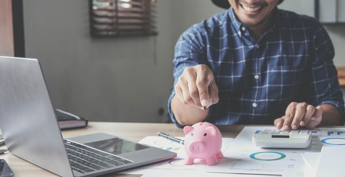 Portrait of an Asian businessman using a calculator to calculate his savings from SME operations, with a pink piggy bank as keep money concept..