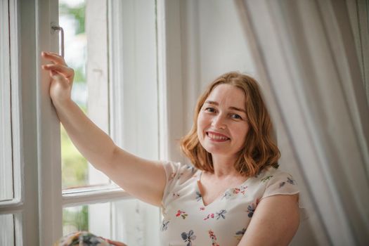 A middle-aged woman in a cream dress sits mysteriously and looks out the window on the windowsill. Green trees outside