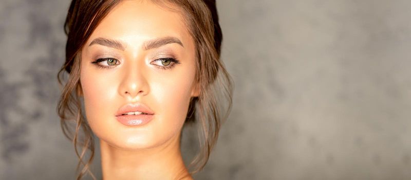 The fashionable young woman. Portrait of the beautiful female model with long hair and makeup. Beauty young caucasian woman with a brown curly hairstyle on the background of a gray wall