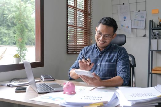 Portrait of an Asian businessman using a calculator to calculate his savings from SME operations, with a pink piggy bank as keep money concept..