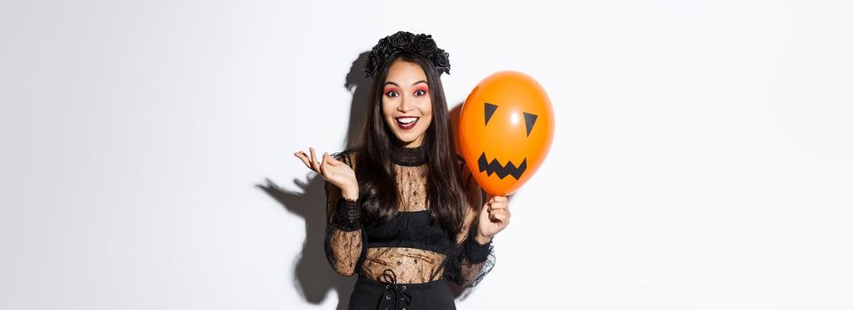 Image of beautiful asian woman celebrating halloween, wearing witch costume and gothic makeup, talking with orange balloon.