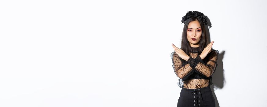 Confident beautiful woman in black gothic dress showing cross gesture, disapprove and stop something bad, disagree with someone about halloween, standing over white background.
