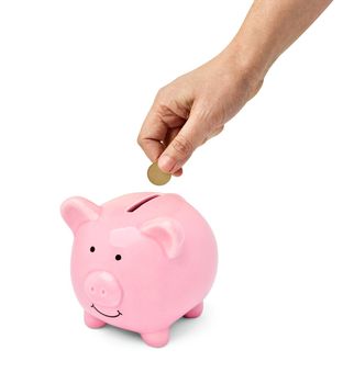 Close up of a hand holding a coin and putting in a pink piggy bank on white background
