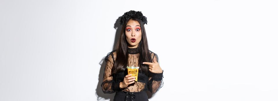 Amazed asian girl looking at camera while pointing finger at sweets, gather treats on halloween, wearing witch dress, standing over white background.