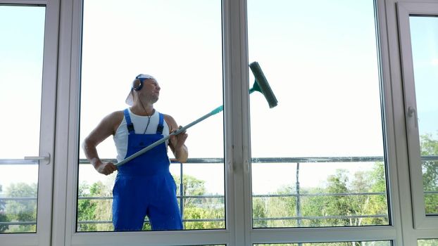 view through window,handsome male worker of cleaning service, in blue overalls, with headphones, , Cleaning, washing Windows by special mop, enjoying his work and music. High quality photo