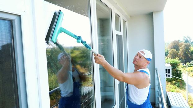 Caucasian handsome man, worker of cleaning service, in blue overalls and in a cap, blaser, Cleaning, washing Windows by special mop, enjoying his work. High quality photo