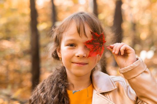Close portrait of a little girl. She covers her eye with a maple autumn leaf. Fall season and children concept