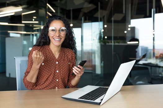 Portrait of successful Latin American business woman, worker in glasses and curly hair looking at camera and smiling happy celebrating triumph and winning success sitting at table holding phone.