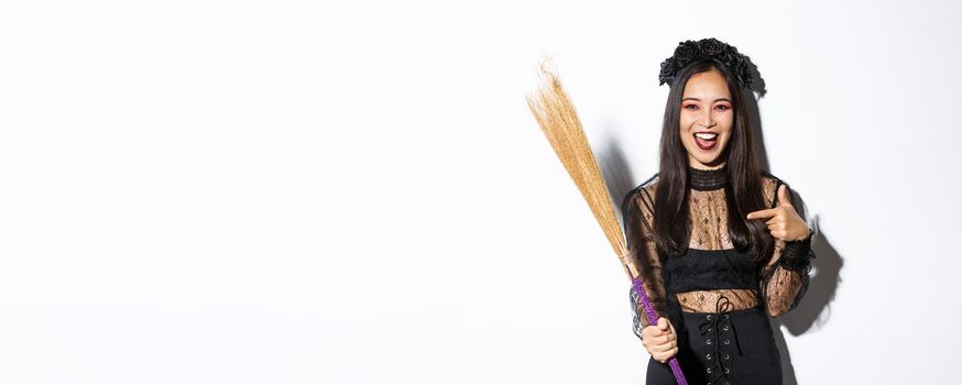 Portrait of attractive woman in witch costume celebrating halloween, pointing at broom, standing over white background.