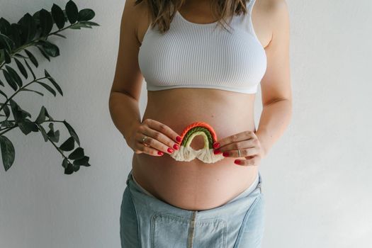 A pregnant girl in a denim jumpsuit holds a rainbow in her hands for the decor of a children's room. Great time to have a baby. Future mom. Rainbow macrame.