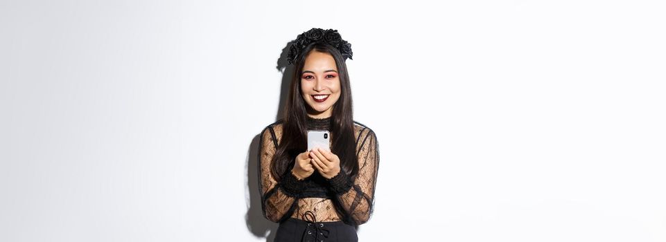 Image of elegant pretty brunette witch in gothic lace dress using mobile phone, smiling at camera, standing over white background.