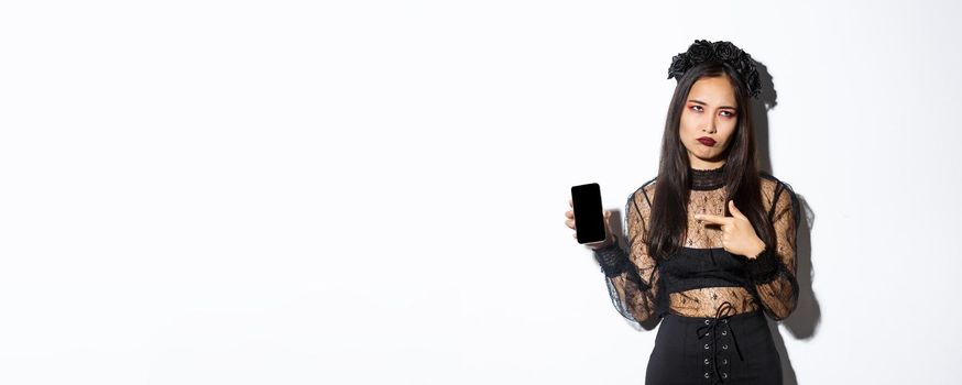 Image of annoyed and reluctant asian girl in witch costume, roll eyes and pouting while pointing finger at mobile phone screen, standing over white background.