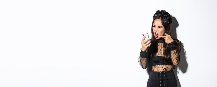 Portrait of angry asian woman in witch costume arguing on video call, shouting at mobile phone and pointing finger, accusing someone, standing over white background.