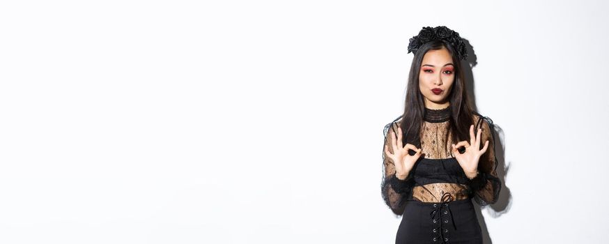 Image of stylish asian woman in gothic dress, looking satisfied and showing okay gestures in approval, praise something good, standing impressed over white background.