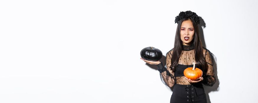 Portrait of disappointed frowning girl in witch costume, holding pumpkins and complaining, grimacing while standing over white background.