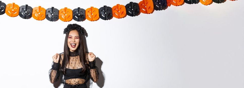 Carefree smiling asian woman in witch costume enjoying halloween party, dancing and rejoicing, standing over white background with pumpkin streamers decoration.