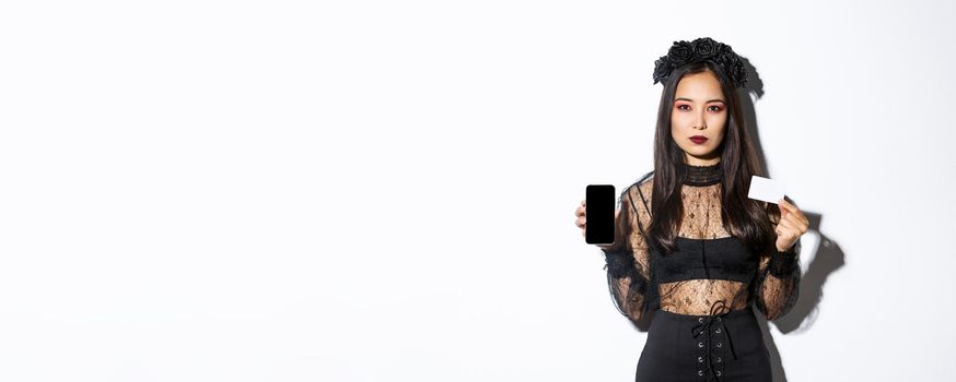 Serious-looking elegant young asian woman in gothic dress and black wreath showing credit card and mobile phone screen, celebrating halloween.