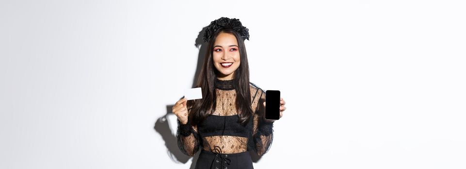Dreamy smiling girl looking away while thinking, showing credit card and mobile phone, wearing halloween gothic dress.
