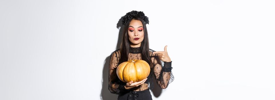 Image of beautiful asian woman in gothic lace dress and black wreath pointing finger at big pumpkin, celebrating halloween, standing over white background.