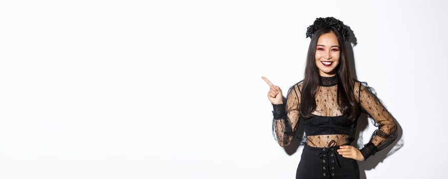 Image of beautiful asian girl in evil witch costume pointing finger upper left corner, showing something for halloween. Woman inviting trick or treat, wearing black lace dress.