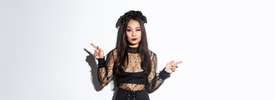 Sassy young evil witch with gothic makeup and wreath, looking arrogant while pointing fingers sideways, showing two halloween themed banners, standing over white background.